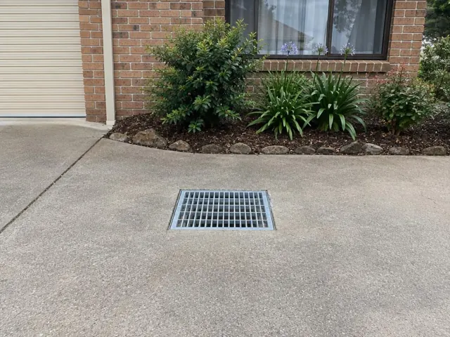 Concrete driveway with a flush stormwater grate beside a brick house and garden bed in Penrith